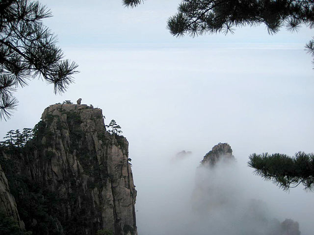Monkey in the distance sitting on top of a prominent rock surrounded by clouds.